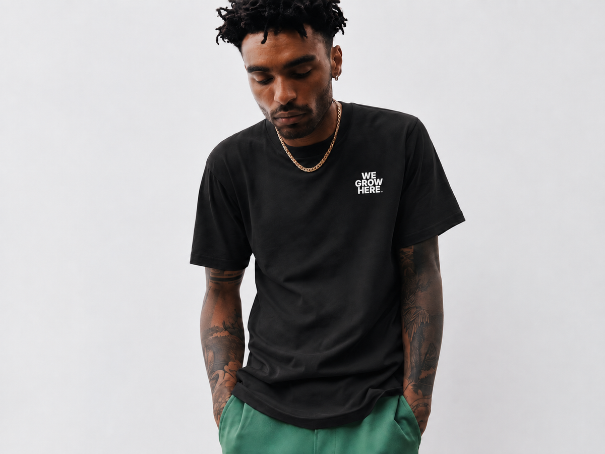 A man wearing a black crewneck t-shirt with the white text 'WE GROW HERE' printed on the left chest, paired with green pants, against a plain light gray background.